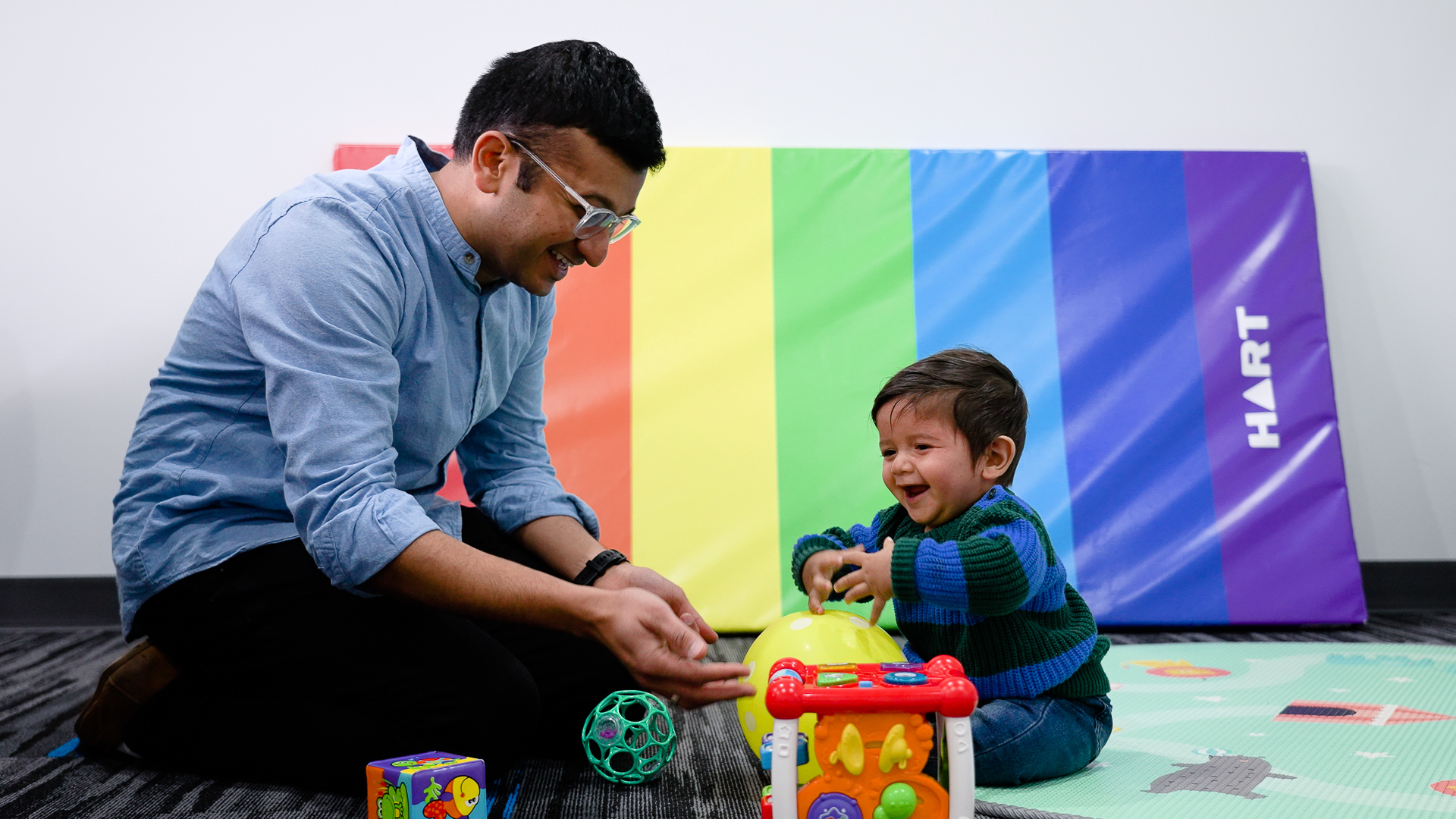 Vishwas Sundaram playing with a 8 month old baby. There is a rainbow coloured playmat in the background and they are surrounded by baby toys
